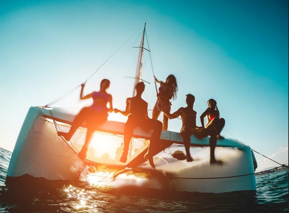 Cinq personnes en maillot de bain ou décontracté, assises et debout sur le pont d'un voilier au coucher du soleil, profitant d'une sortie en mer.