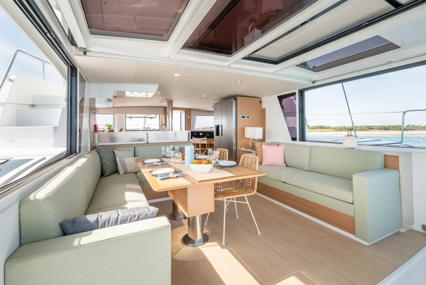 Intérieur d'un salon d'une bateau avec tables, canapés et vue sur l'eau par les fenêtres.