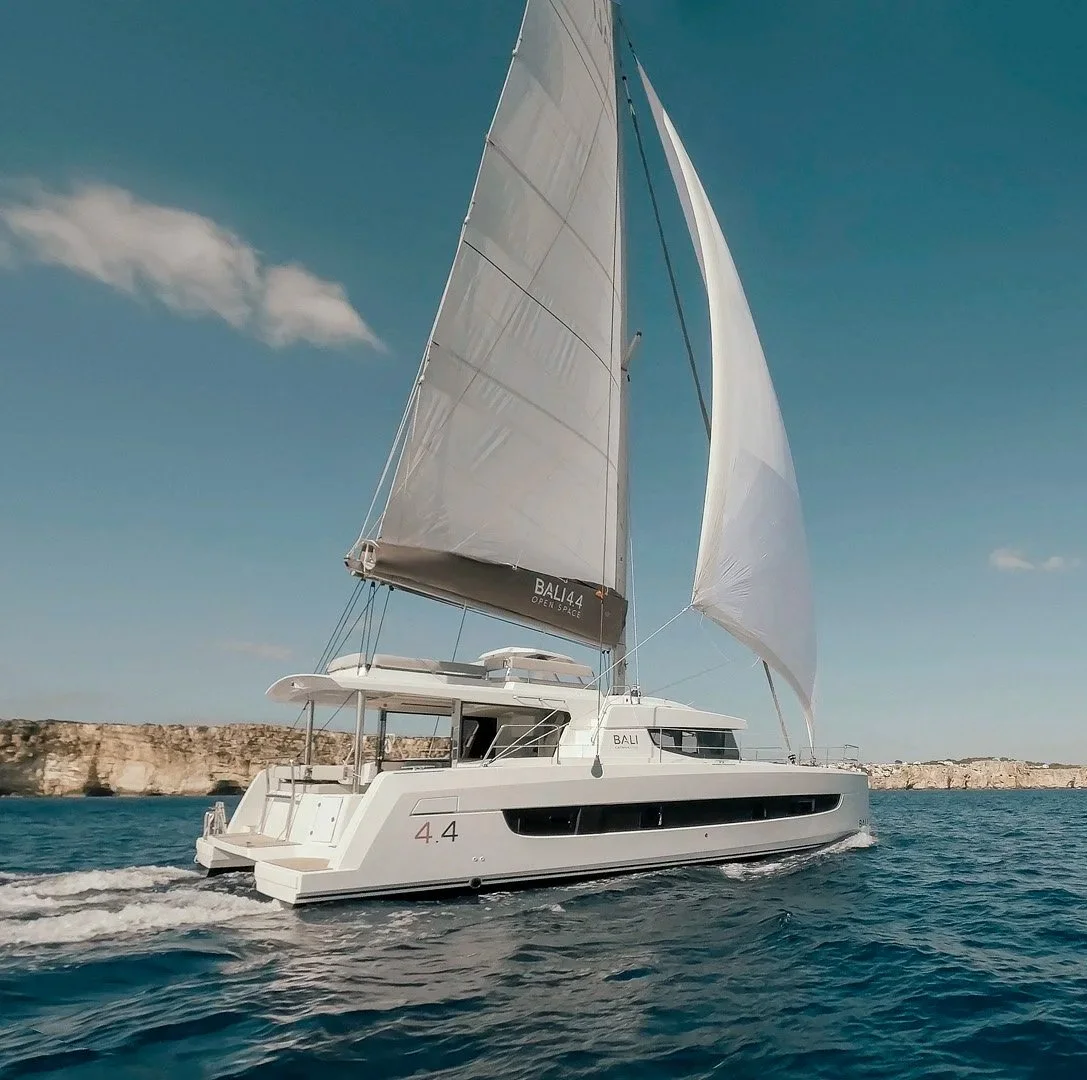 A white sailboat with large sails moving across blue water near a rocky coastline under a partly cloudy sky.