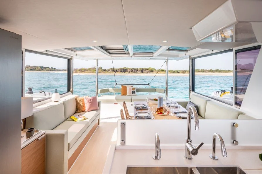 Intérieur d'un yacht avec vue sur l'eau, salon moderne avec canapé blanc, table à manger avec assiettes, verres et nourriture, grandes fenêtres offrant une vue panoramique.
