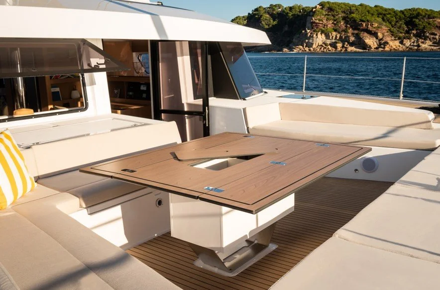 Une vue de la terrasse d'un yacht avec un mobilier en blanc, incluant un grand coussin beige, et une table en bois avec un hayon central ouvert, face à la mer et à un paysage rocheux verdoyant.