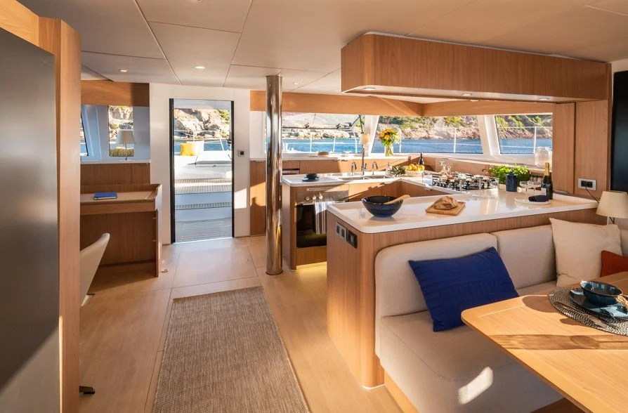 Intérieur d'une cuisine moderne à bord d'un bateau, avec vue sur l'eau, comptoir en marbre, îlot central, vaisselle, plantes et fleurs, murs en bois clair et grand éclairage naturel.