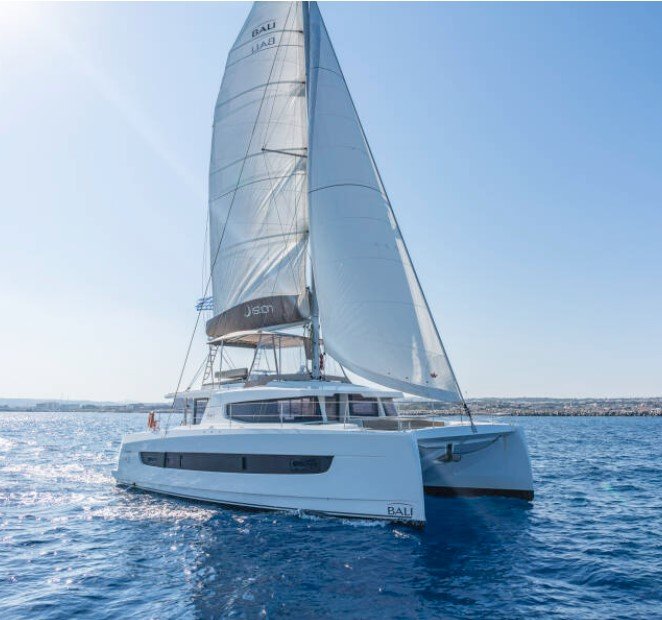 A large white sailing catamaran with sails open on a calm body of water under a clear blue sky.