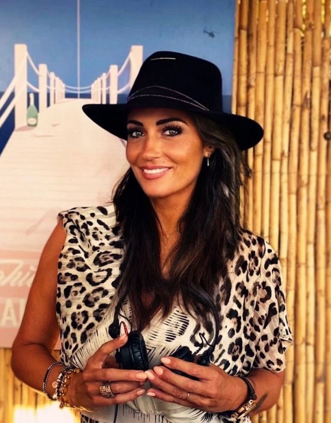 Une femme souriante portant un chapeau noir et une robe à motif léopard, tenant des écouteurs, dans un décor avec un panneau en arrière-plan et un mur en bambou.