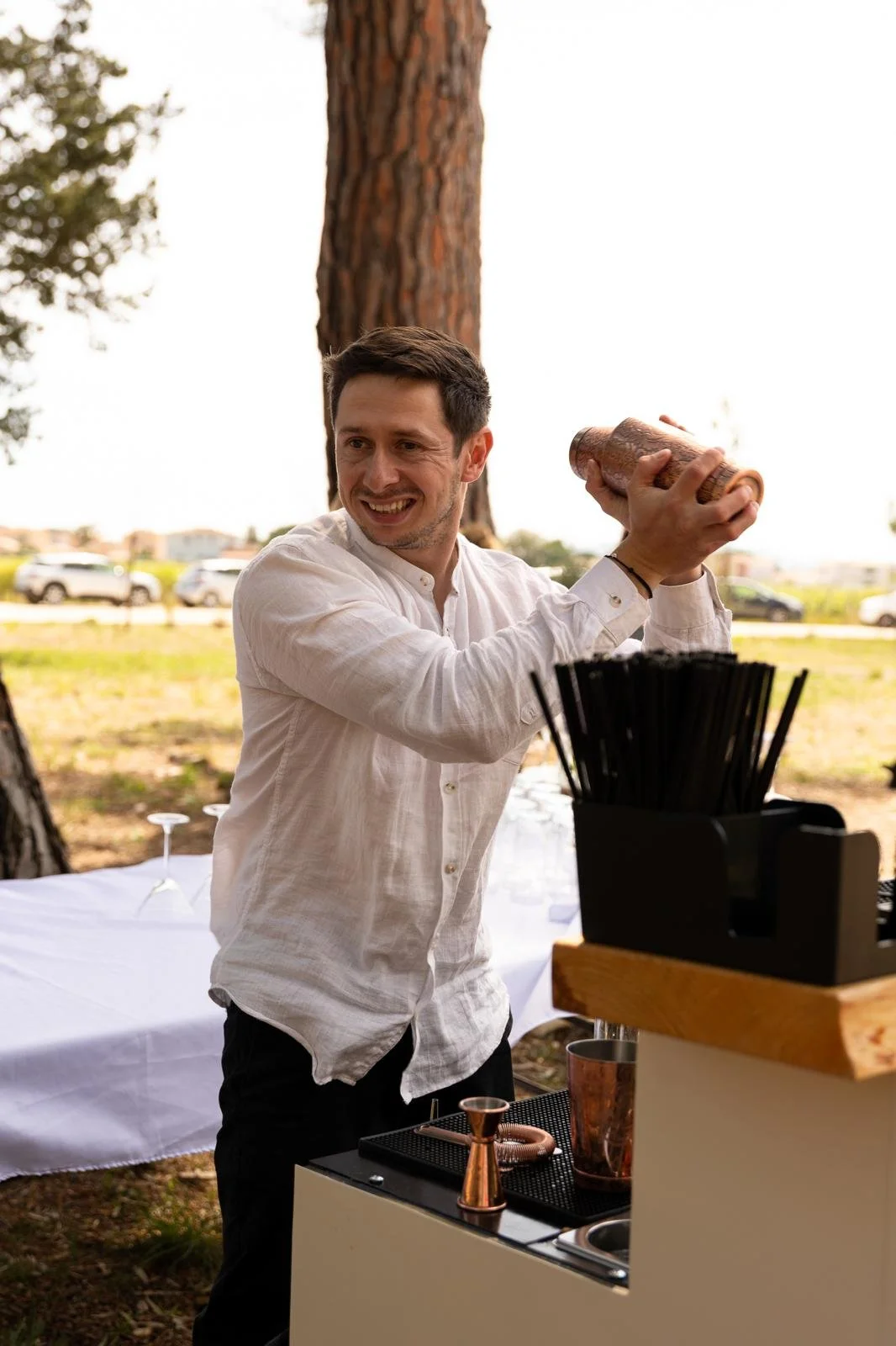 A smiling man in a white shirt preparing a drink outdoors, with trees and cars in the background.