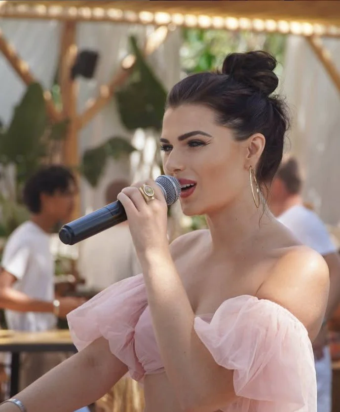 A woman with dark hair styled in a loose updo, wearing large hoop earrings and a pink off-the-shoulder top, singing into a microphone at an outdoor event with blurred background of people and greenery.