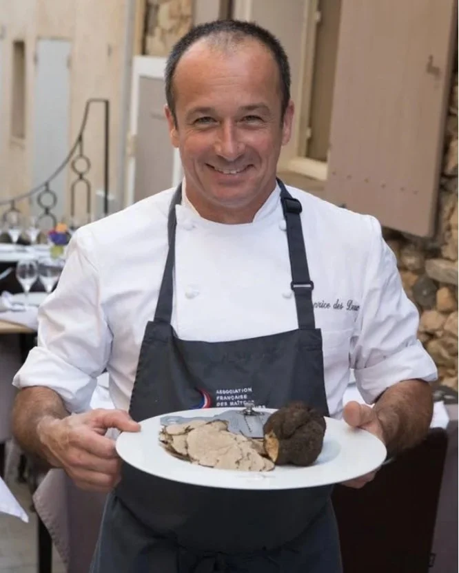 A smiling male chef in a white uniform and black apron holding a plate with pieces of beef and a bread roll, standing in a restaurant setting.