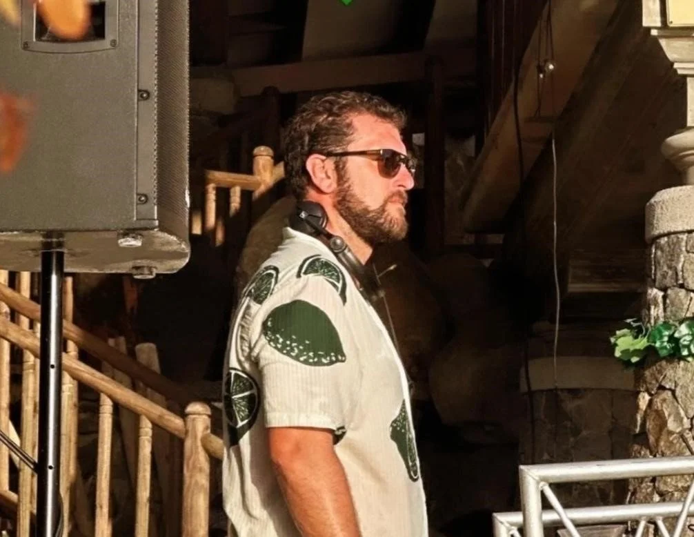 Un homme portant des lunettes de soleil et une chemise imprimée de citrons verts, se tenant en profil dans un intérieur rustique avec des escaliers en bois et un mur en pierre. Italian DJ