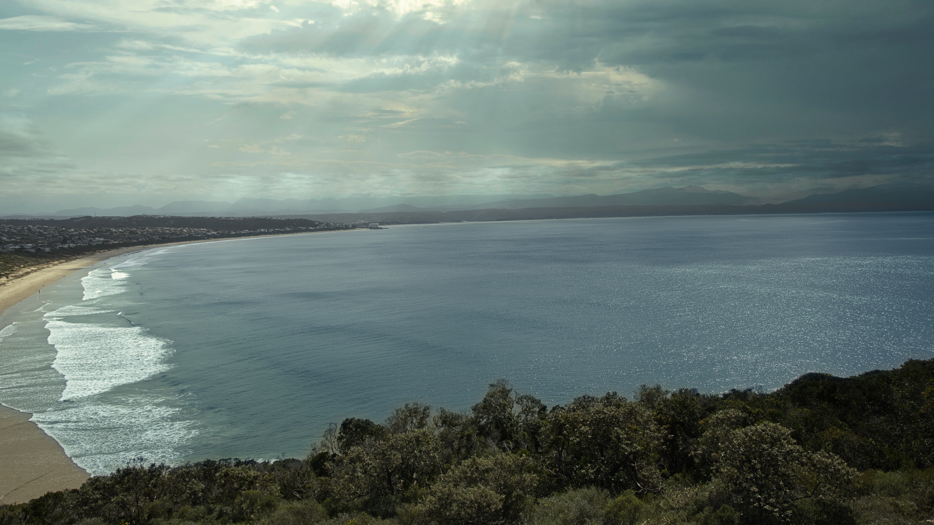 Aerial view of Plettenburg Bay, coastline, and ocean, with hills and a cloudy sky in the distance.
