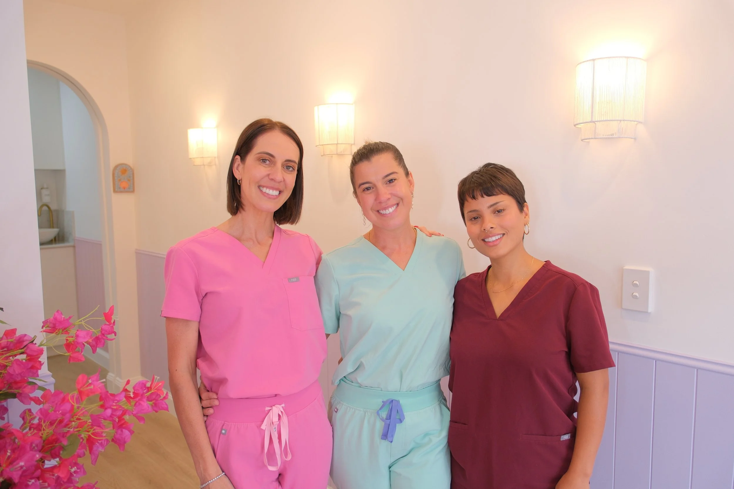 Three smiling women in colorful medical scrubs standing close together indoors, with a white wall, wall lamps, and pink flowers visible in the background.
