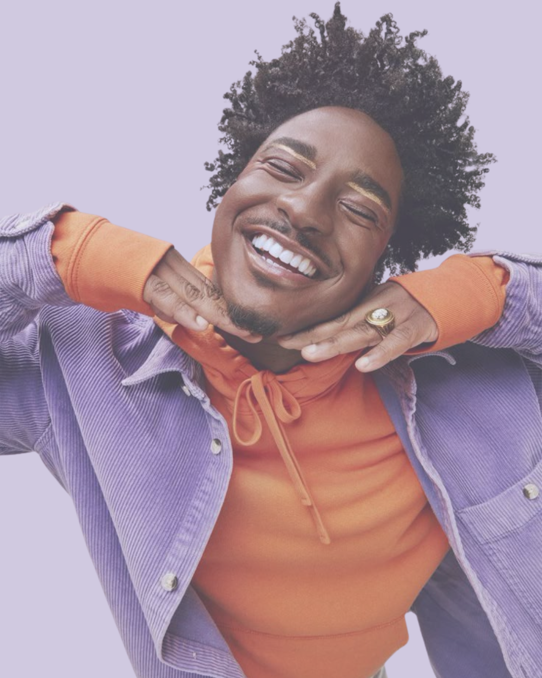 A joyful person with curly hair, smiling widely with eyes closed, wearing an orange hoodie and a purple jacket, with hands resting under chin, against a light purple background.