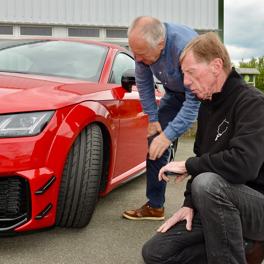 Zwei ältere Männer besprechen ein rotes Auto im Freien.