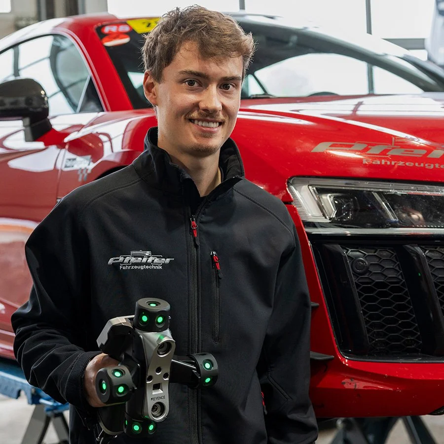 Ein junger männlicher Techniker steht vor einem roten Auto, hält ein hochentwickeltes Messgerät, trägt eine schwarze Jacke mit Firmenlogo, im Hintergrund einen Auto-Reparaturraum.