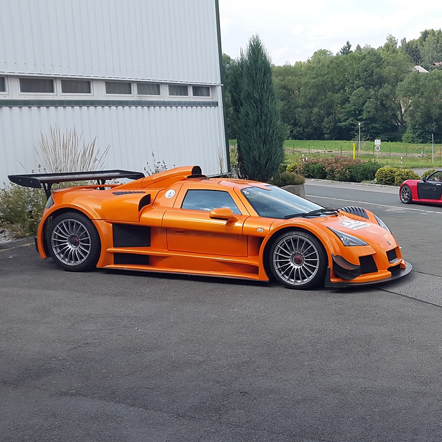 Ein orangefarbener Rennwagen parkt auf einem Parkplatz neben einem weißen Gebäude, im Hintergrund Bäume und ein weiteres Auto.