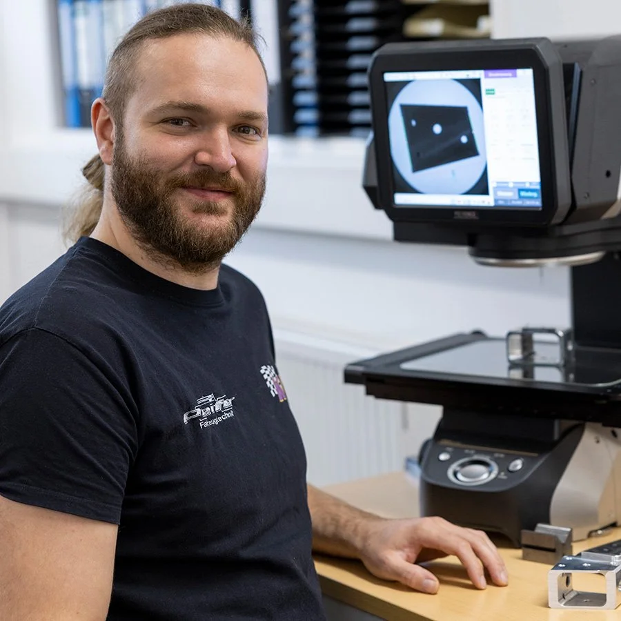 Ein Mann mit Bart sitzt an einem Tisch mit elektronischer Mess- und Prüftechnik, während er die Kamera des Geräts bedient. Im Hintergrund ist ein Bildschirm mit einer Diashow der Messdaten zu sehen.