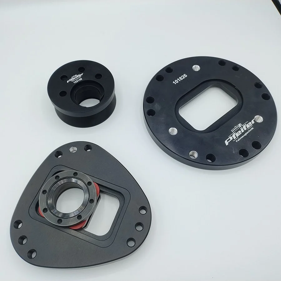 Drei schwarze technische Komponenten auf weißem Hintergrund, eine mit Kreisbohrungen, eine mit rechteckiger Öffnung und ein Merkmal mit rot gearbeiteten Elementen.