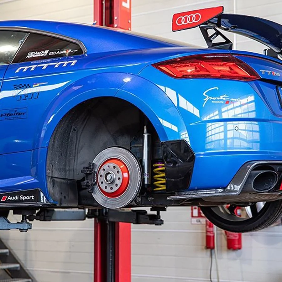 Ein blauer Rennwagen auf einer Hebebühne, keine Felge und Reifen montiert, sichtbare Bremsscheibe, Spoiler im Heck, im Hintergrund Feuerlöscher.