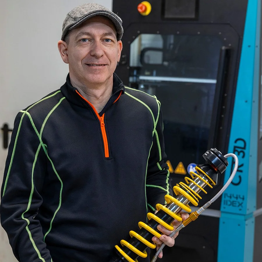 Ein Mann mit grauem Hut und schwarzen Jacke mit neon grünen Nähten hält ein gelbes Fahrzeugelement, wahrscheinlich eine Stoßdämpfer, in der Hand.