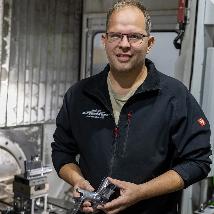 Ein Mann trägt eine schwarze Jacke mit Firmenlogo und hält ein Metallteil in den Händen, im Hintergrund eine Werkstatt mit Maschinen.