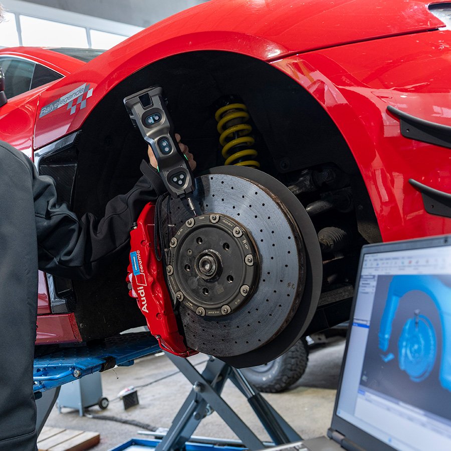 Ein rotes Auto auf einer Hebebeschraube, mit offenem Rad, geöffneter Bremszange und sichtender Radnabe, während ein Techniker es repariert oder wartet, mit einem Laptop im Vordergrund, der eine digitale 3D-Darstellung des Autos zeigt.