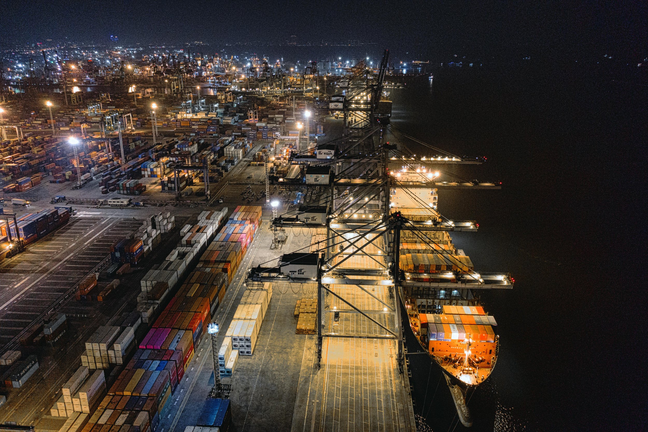 Nighttime view of a busy shipping port with containers, cranes, and a container ship docked at the pier.