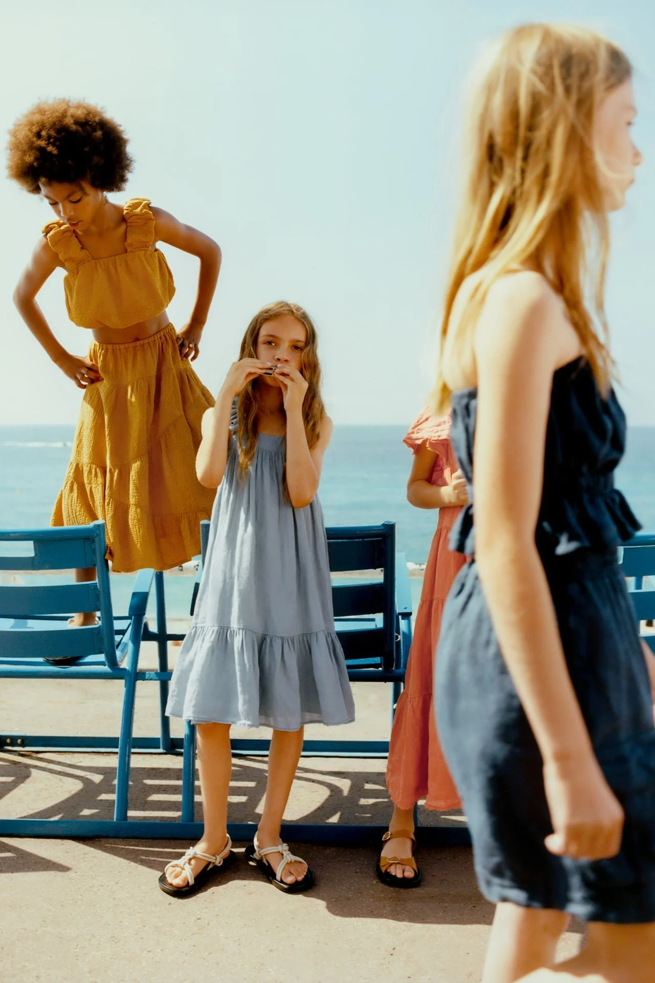 Four young girls standing near blue chairs on a beach, with the ocean in the background. One girl with long blonde hair does not face the camera. Another girl with red hair and a blue dress is in the center, holding her hands near her mouth. A girl w