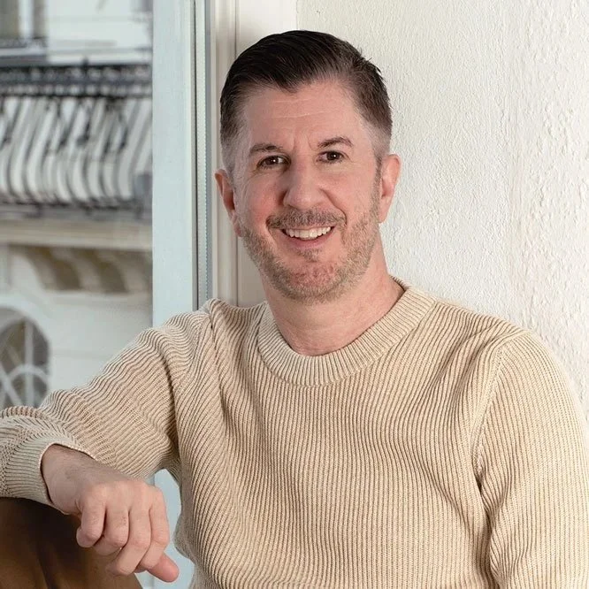 Ein lächelnder Mann in einem beige Pullover sitzt vor einem Fenster, im Hintergrund sind Geschirr und Geschirrtücher sichtbar.