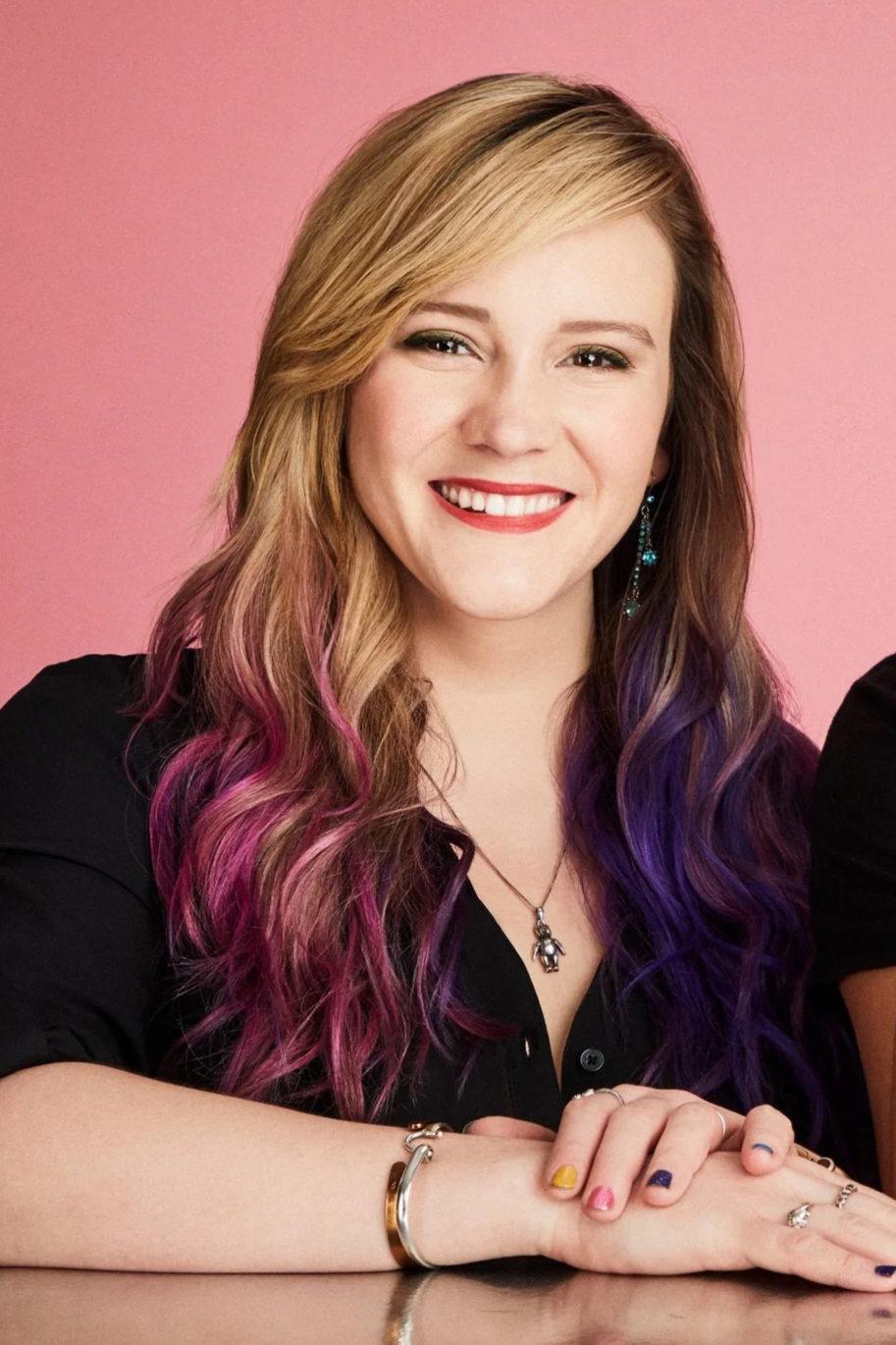A young woman with long, wavy hair that transitions from blonde to pink and purple, smiling, wearing a black shirt, colorful nail polish, jewelry, and earrings, with a pink background.