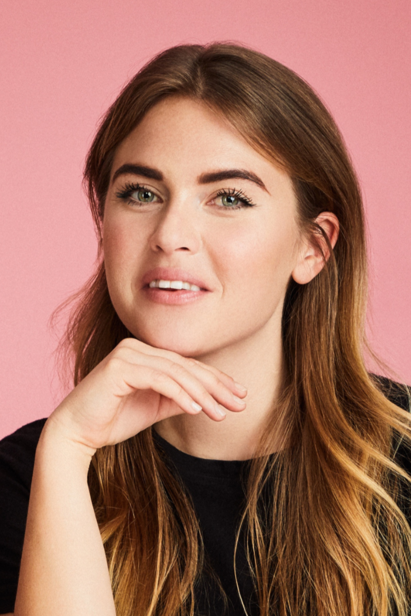 A woman with long brown hair and green eyes, resting her chin on her hand, against a pink background.