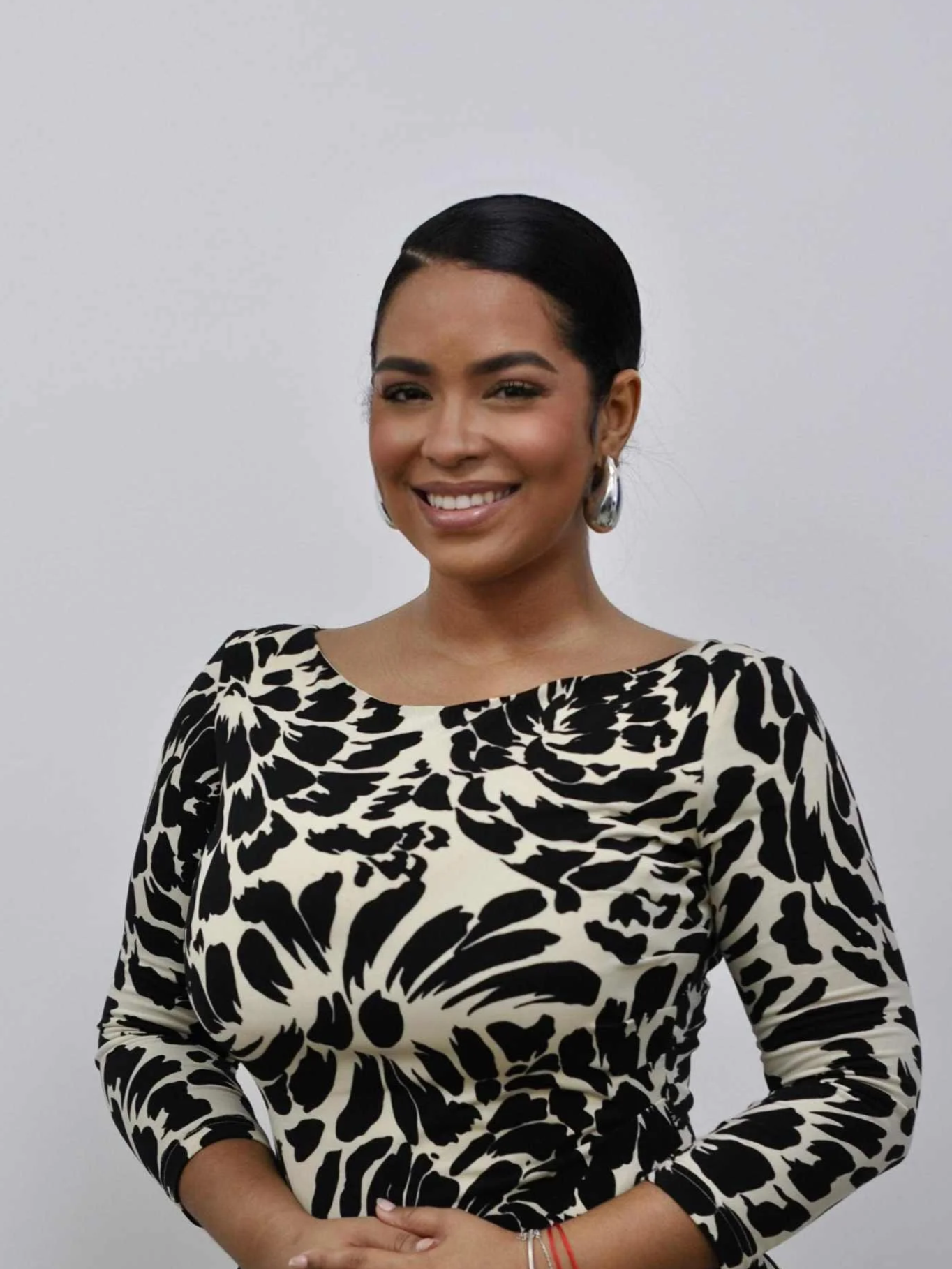 A woman with styled black hair, wearing a black and white floral patterned top, smiling, and standing against a plain white background.