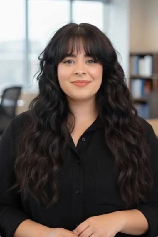 A woman with long, wavy dark hair wearing a black button-up shirt, smiling in an office setting with bookshelves and large windows in the background.