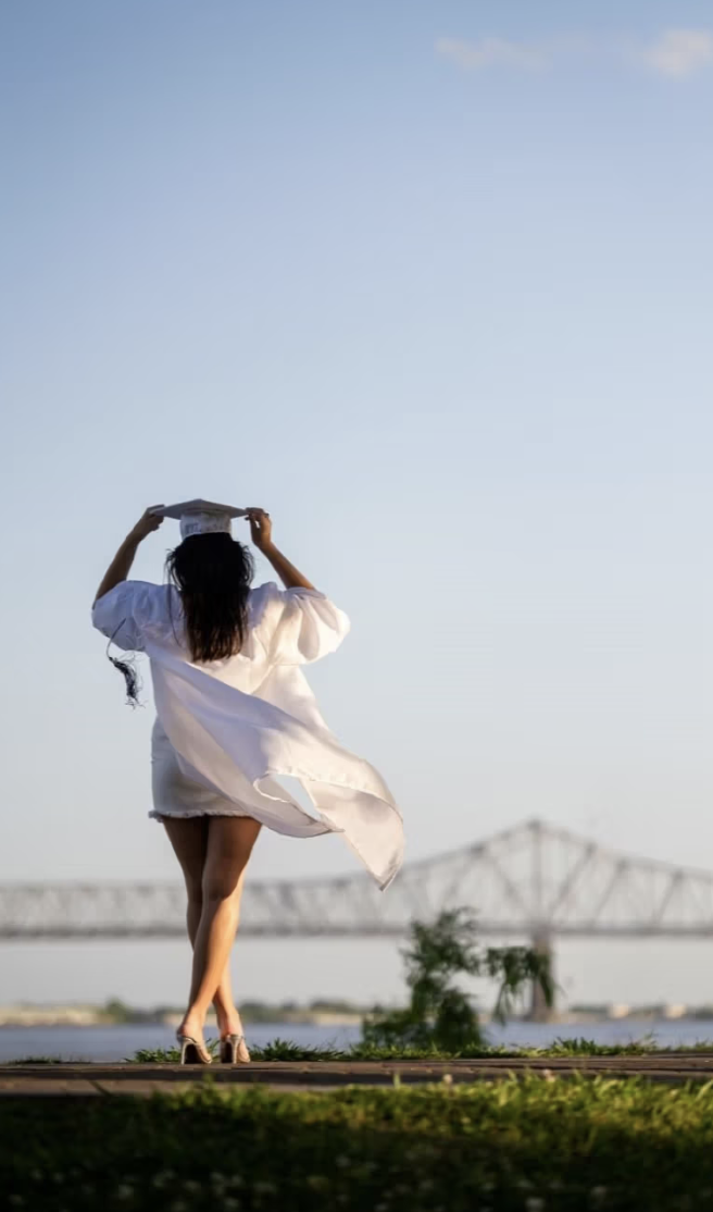 A woman in a white dress holding a diploma above her head outdoors with a bridge in the background.