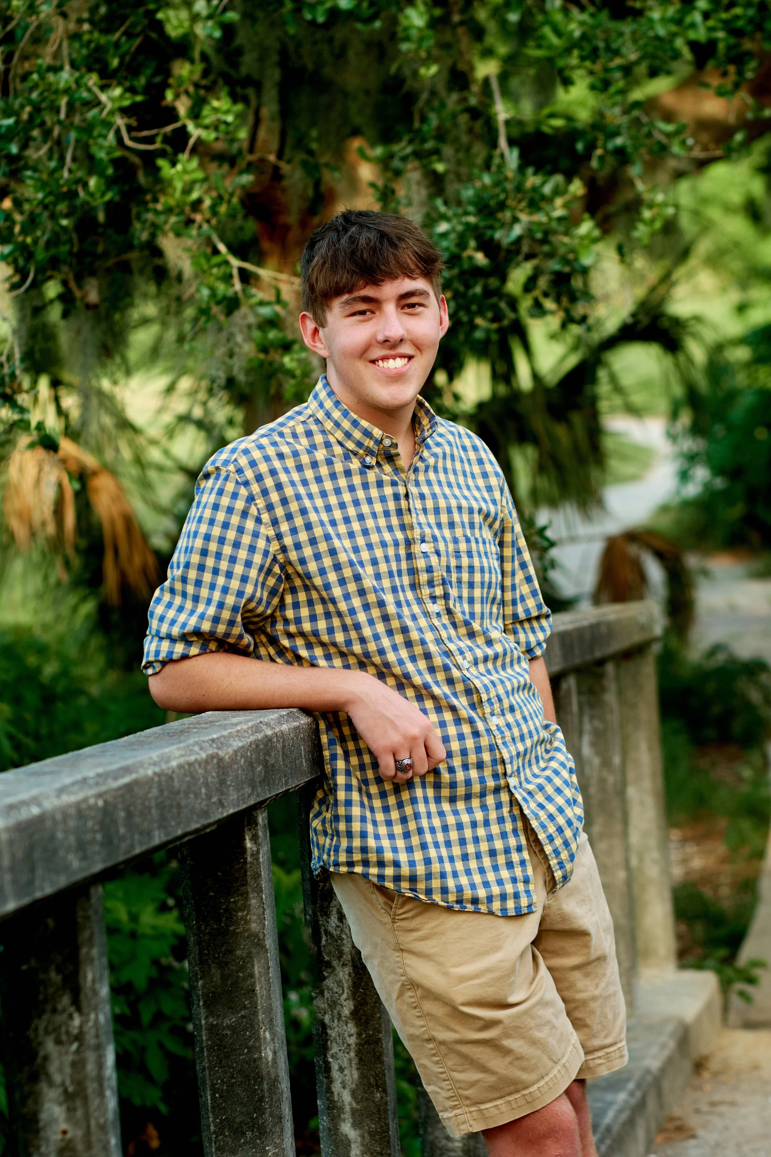 A young man with brown hair, wearing a yellow and blue checkered shirt and tan shorts, leaning against a wooden railing outdoors with greenery in the background.
