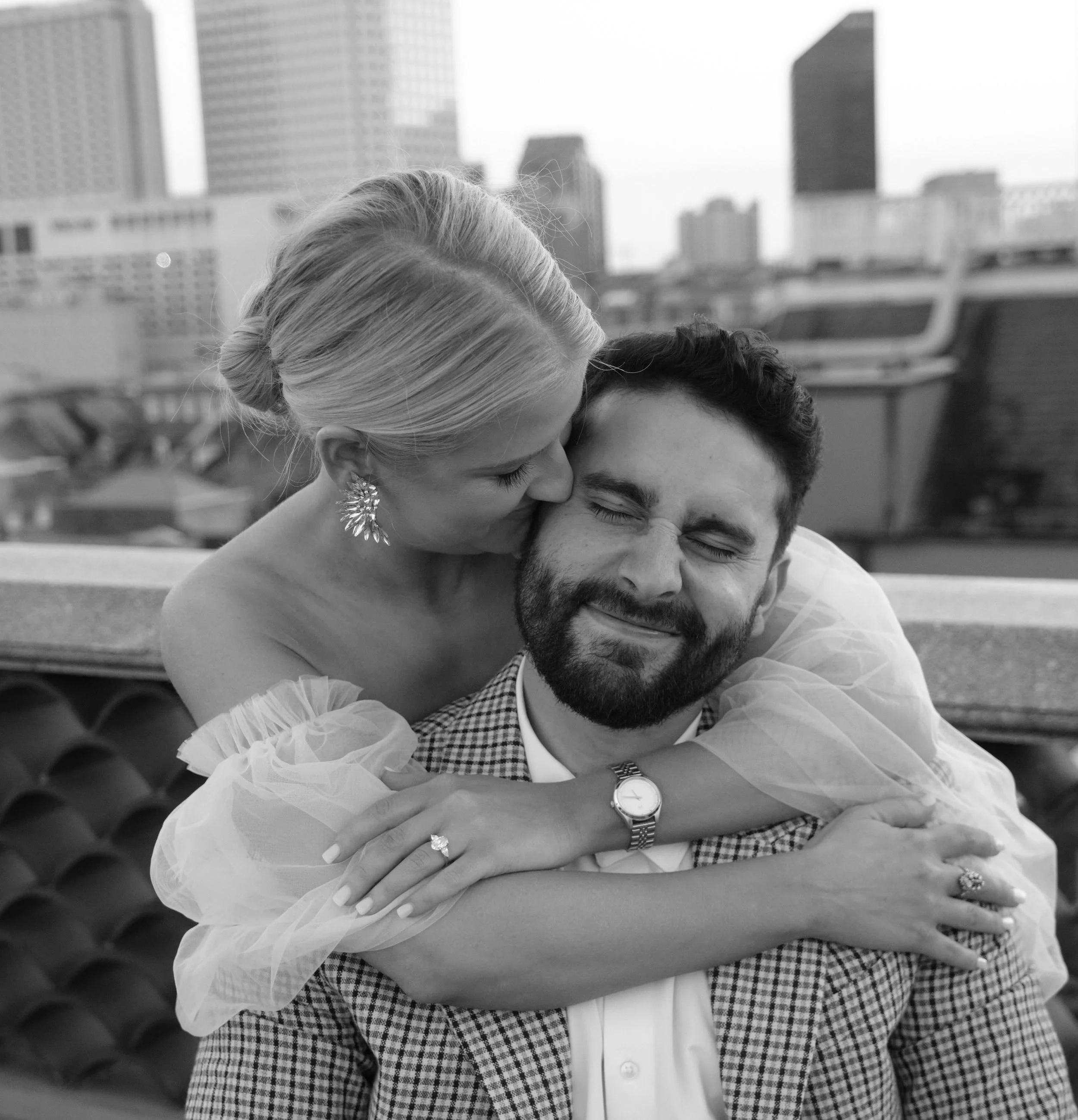A woman kisses a man on the cheek in an outdoor city setting, the man is smiling with eyes closed, and they are embracing each other.