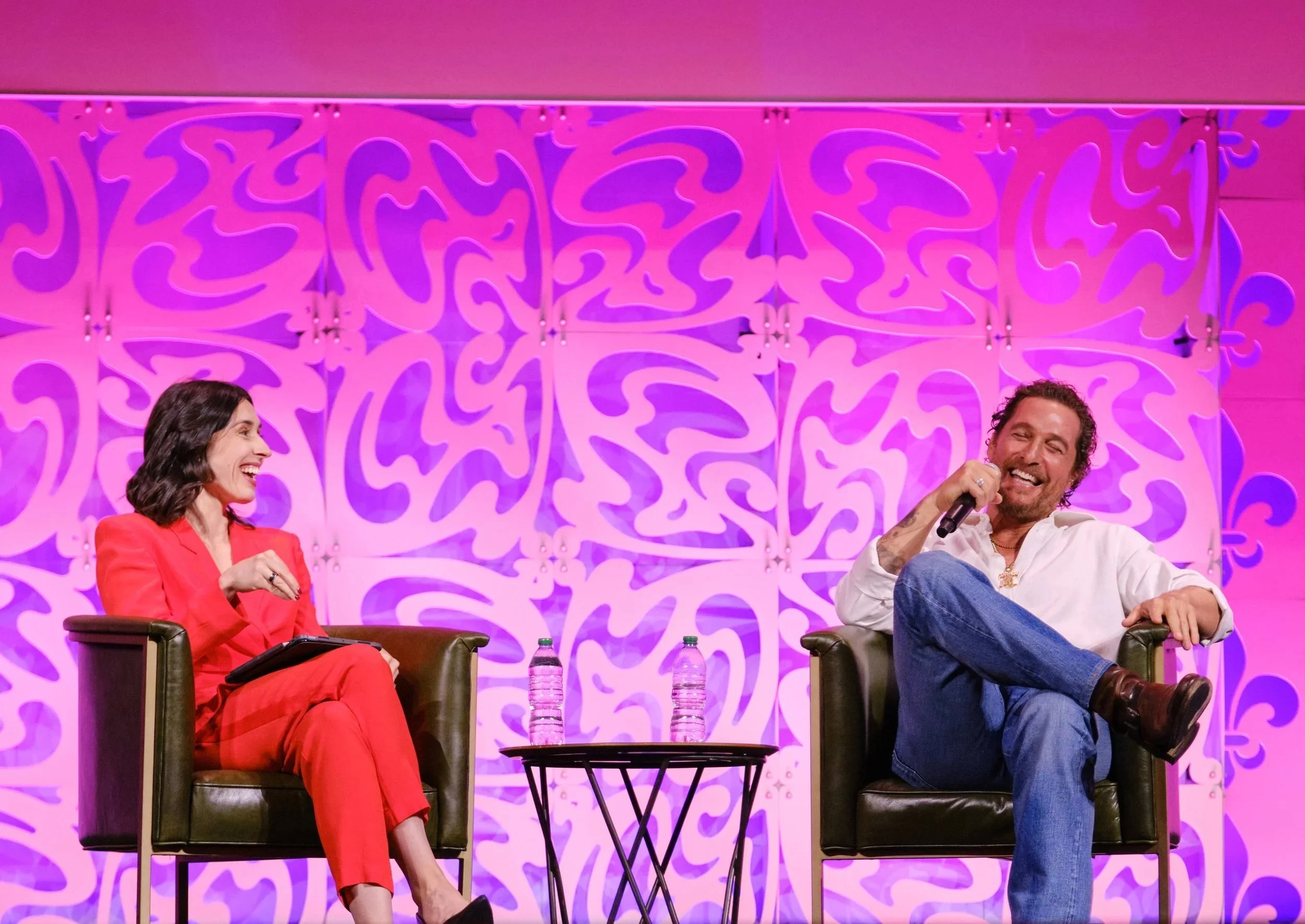 A woman and a man are sitting on stage in black chairs, engaging in a conversation, with a purple decorative backdrop behind them. The woman is wearing a red suit and smiling, while the man, dressed in a white shirt and blue jeans, is holding a micro