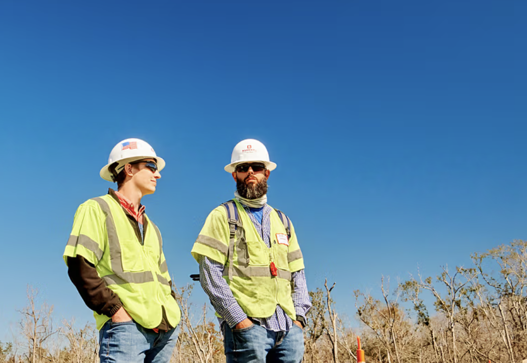 Two construction workers standing outdoors with blue sky behind them, wearing protective helmets and high-visibility safety vests.