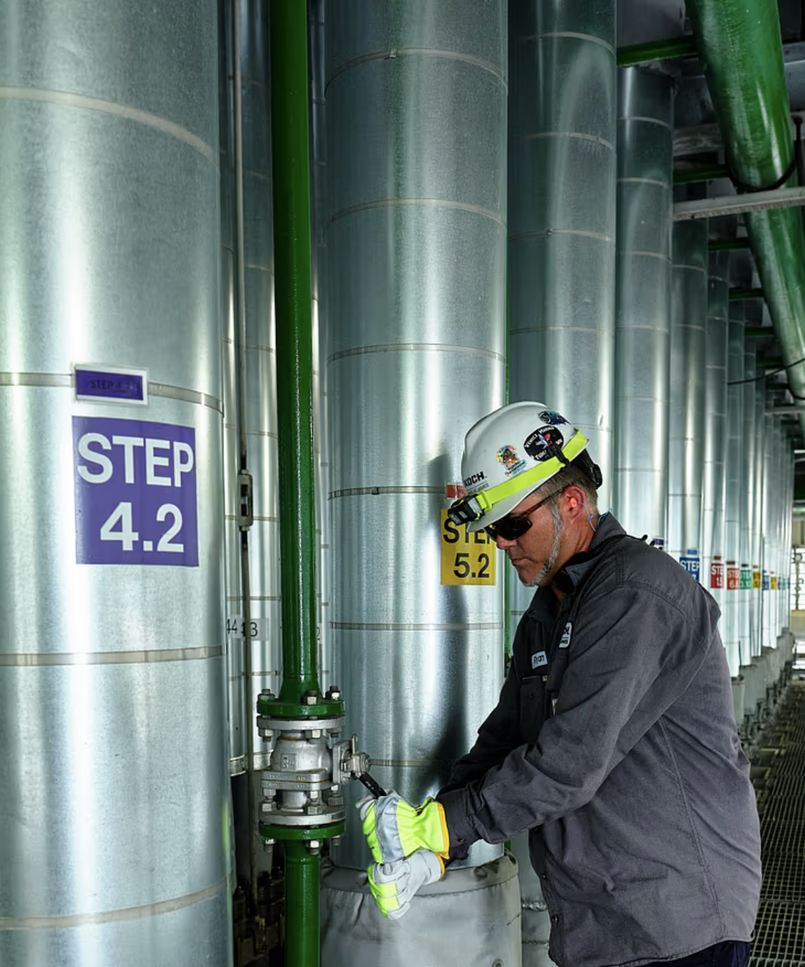 A worker in safety gear, including a white helmet with stickers, sunglasses, and gloves, is working on a green pipe in an industrial plant. Behind him are large silver tanks labeled with step numbers, such as 'STEP 4.2' and 'STEP 5.2.'