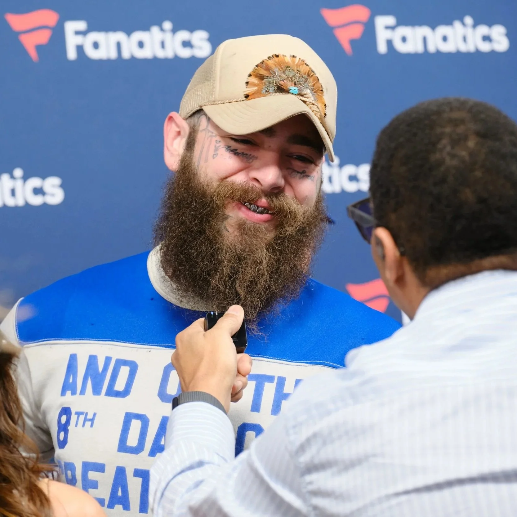 A mpost malone smiling with  tattoos on his face is being interviewed at a Fanatics event, smiling and wearing a beige cap decorated with feathers.