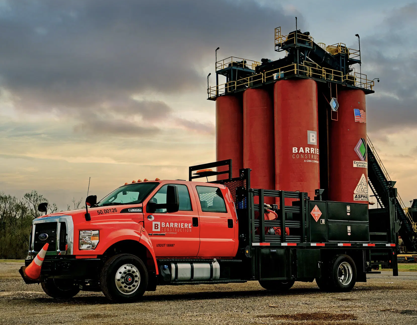 A red Barriere construction truck with a black flatbed parked on an asphalt surface, featuring orange traffic cones hanging on the front, and a large red industrial structure in the background, under a cloudy sky. GK Photography New Orleans