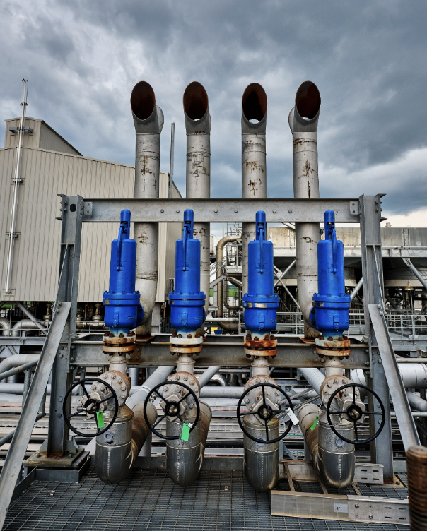 Industrial pipes with blue valves and large exhaust stacks under cloudy sky.