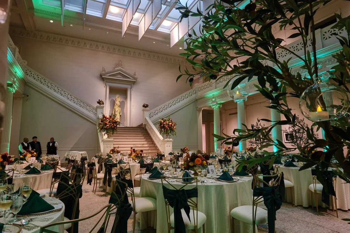 Elegant banquet hall decorated for a wedding reception, with round tables set with white tablecloths, glassware, black napkins, and floral centerpieces. A staircase with floral arrangements leads to a religious statue on a pedestal at the front, and there are green accent lights illuminating the room's classical architecture.