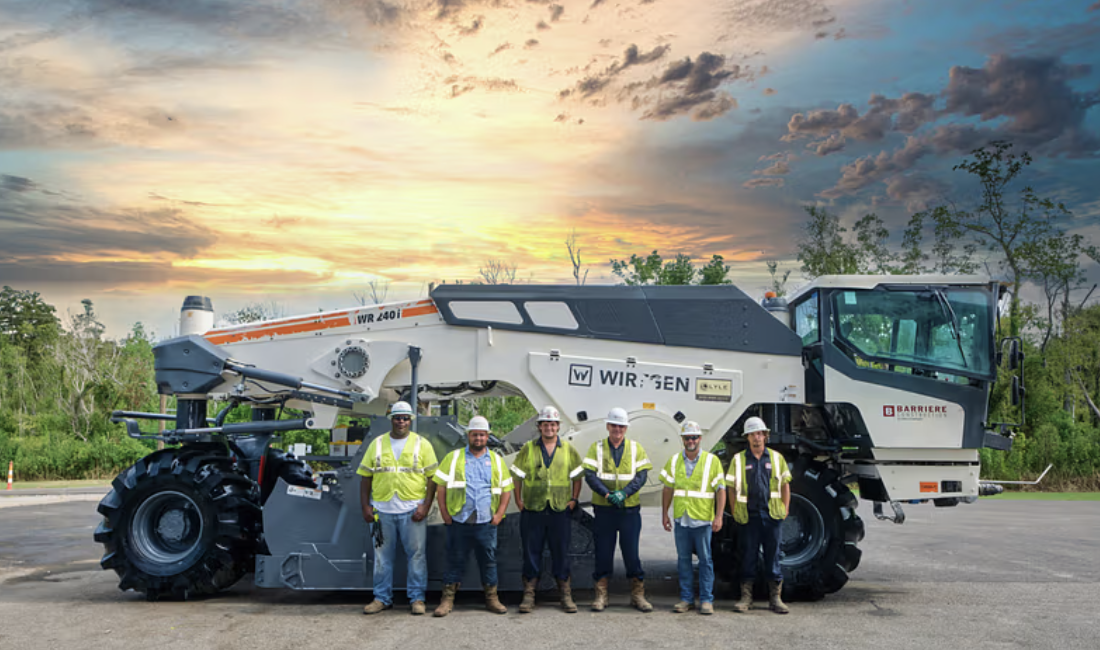 Seven construction workers in safety vests and helmets standing in front of a large road construction machine at sunset.