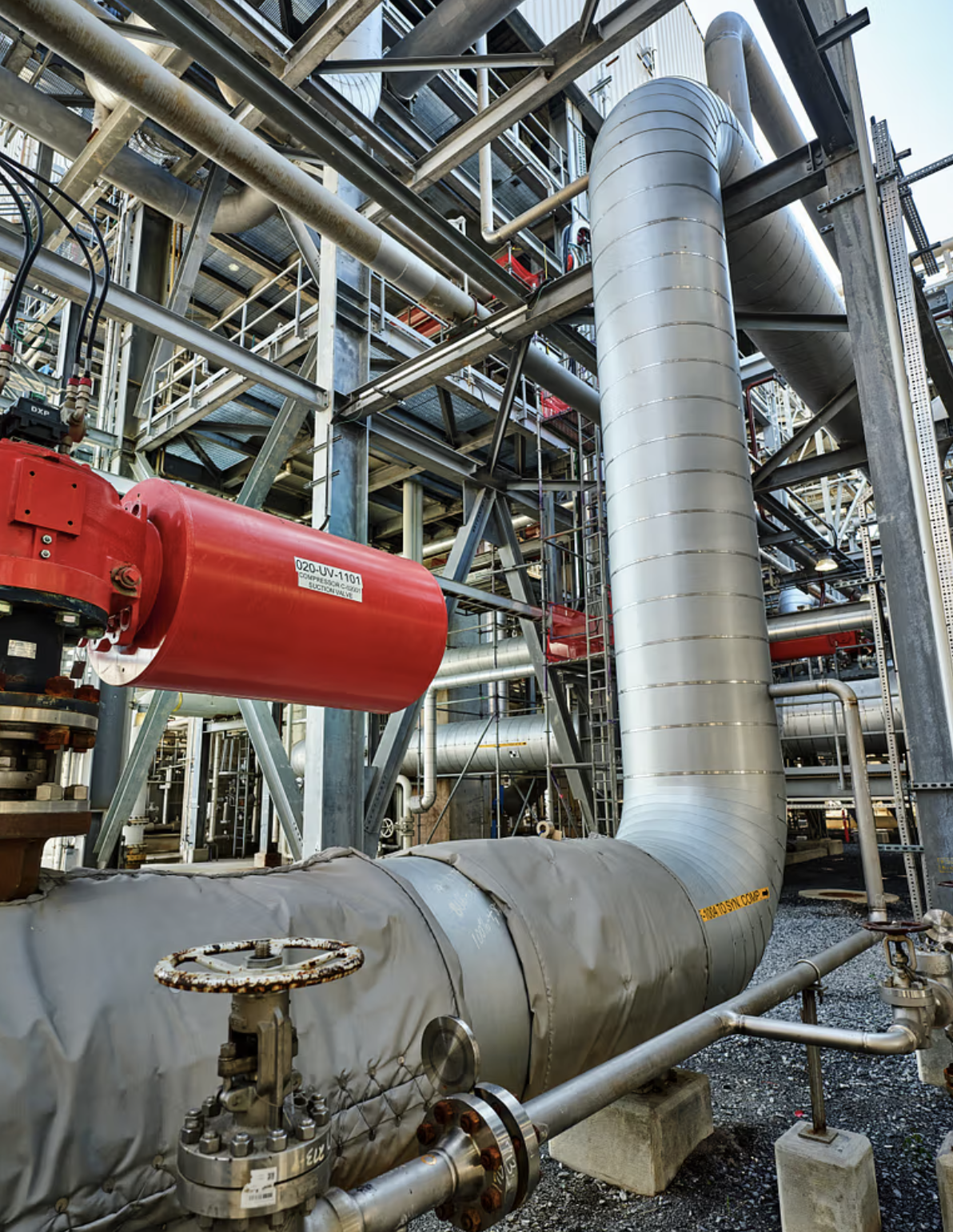 Industrial piping system with large silver and red pipes, valves, and scaffolding in a factory or plant.