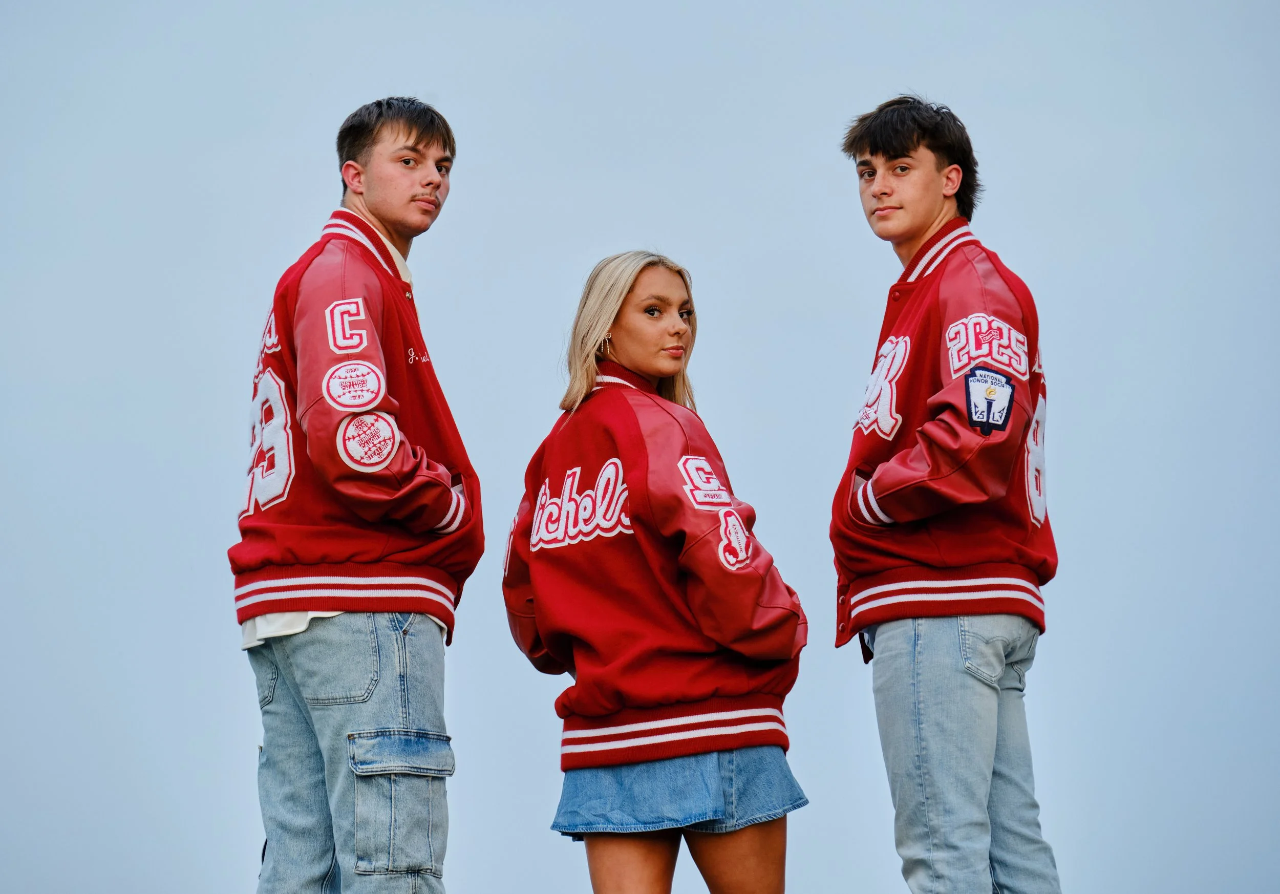 Three young individuals standing against a light blue sky, all wearing red letterman jackets and casual blue jeans or denim skirts, looking directly at the camera with serious expressions.
