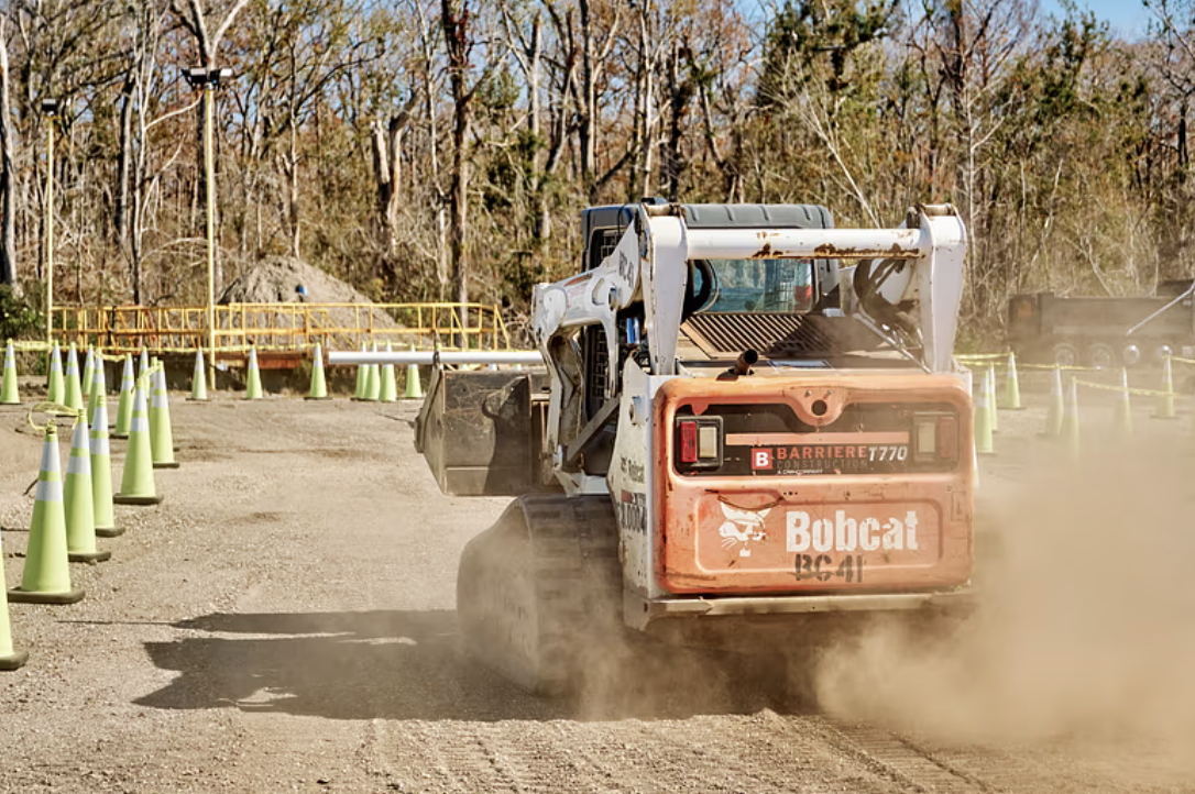 A Bobcat compact loader moving dirt at a construction site surrounded by orange safety cones and trees.