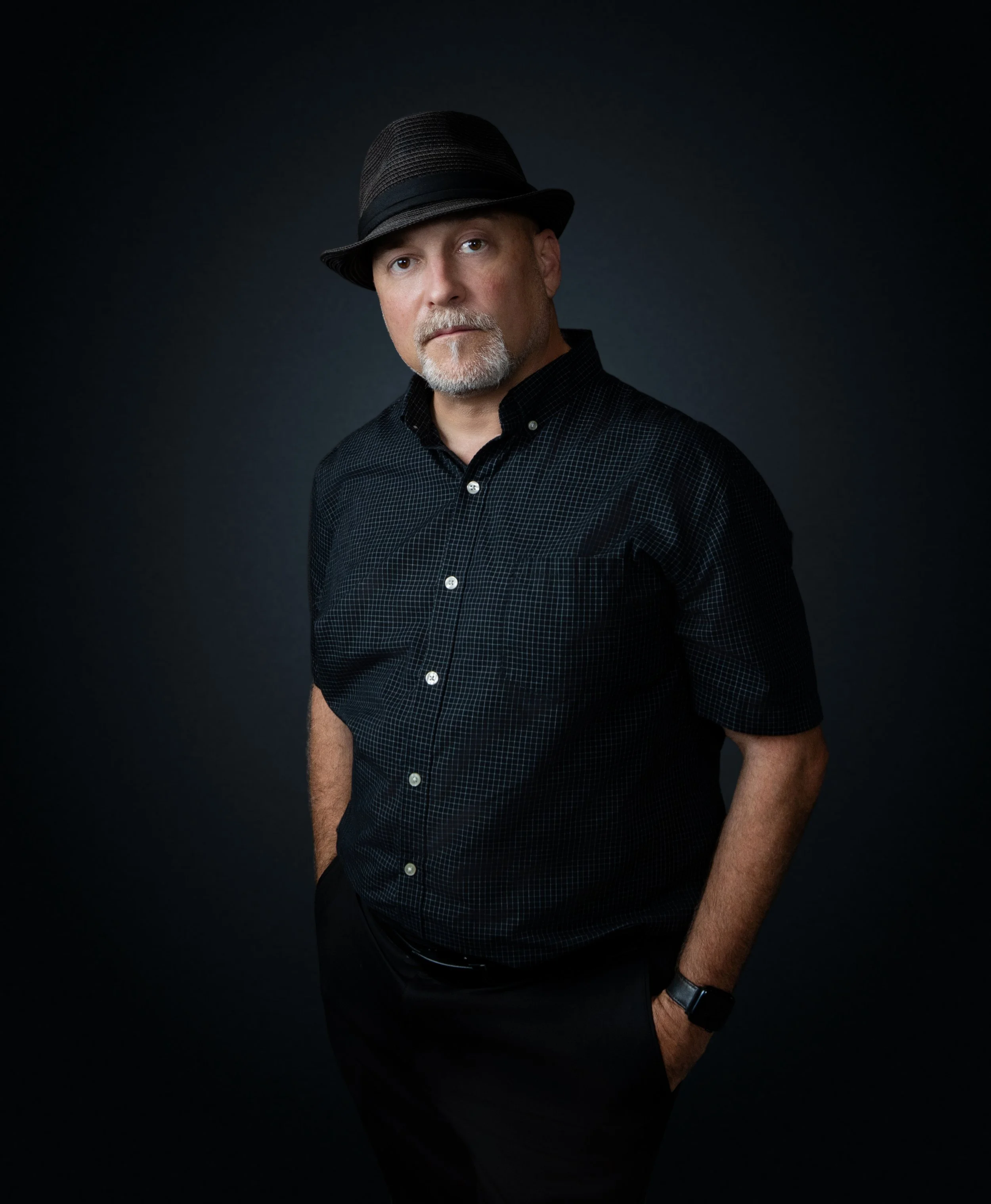A man wearing a black fedora hat, a black checkered shirt, black pants, and a wristwatch, standing against a dark background.