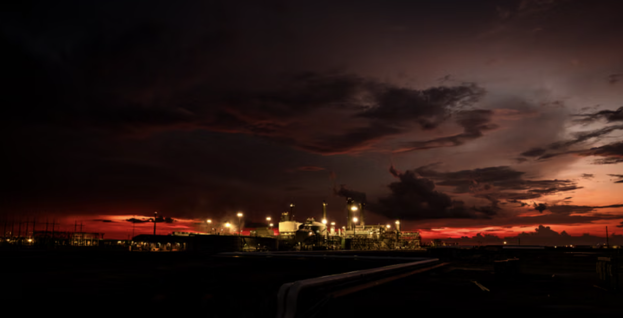 Industrial factory or refinery at dusk with smoke stacks emitting dark smoke and vibrant red and orange sunset sky.