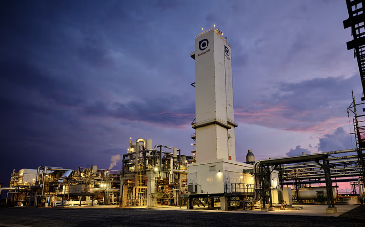 Refinery or industrial plant at dusk with pipes and machinery, and a tall cooling tower or tower structure.