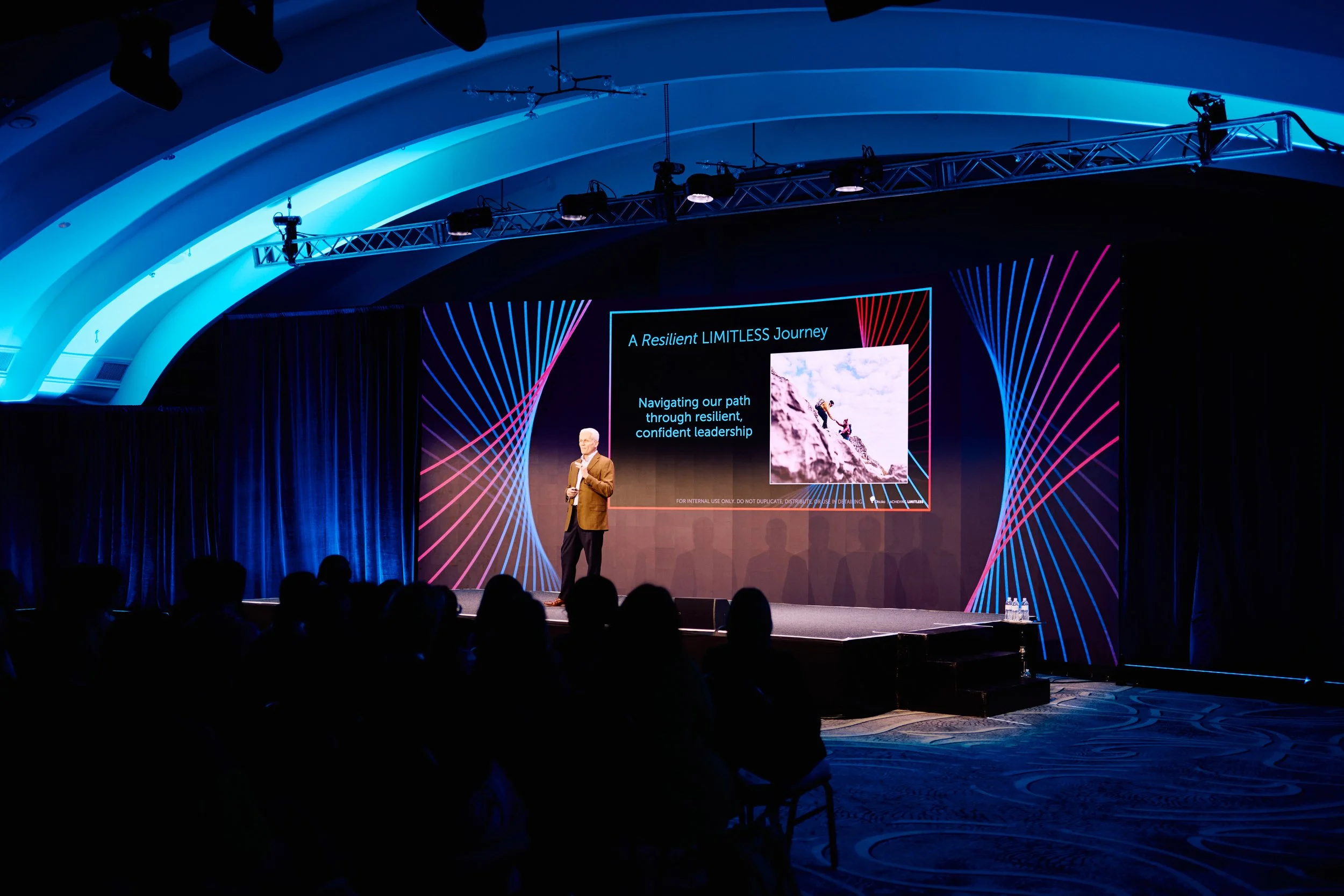 A man giving a presentation on stage with a large screen behind him displaying a slide titled 'A Resilient LIMITLESS Journey.' The slide shows an image of climbers ascending a mountain and text about resilient leadership. The stage is decorated with blue and pink light patterns, and the audience is seated in darkness.