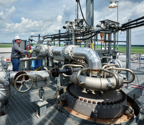 Industrial worker operating large machinery outdoors, with pipes and equipment on a metal platform under a partly cloudy sky.